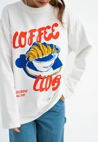 Witte sweatshirt met een kleurrijke grafische print van een croissant en een koffiekop. Tekst leest "COFFEE CLUB" in het rood en blauw. Casual pasvorm.
