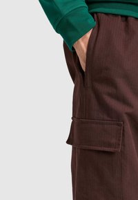 Pantalones cargo marrones con textura acanalada vertical, bolsillos laterales y un bolsillo con solapa. Combinados con una camisa de manga larga verde.