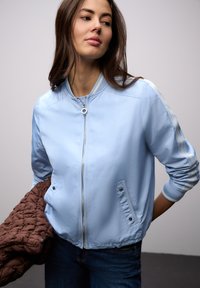 Mujer con chaqueta bomber azul claro con cremallera y vaqueros, sosteniendo una chaqueta marrón matelassé doblada, mirando ligeramente hacia su izquierda.