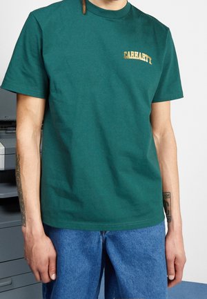Osoba nosząca zielony t-shirt Carhartt z żółtym logo na piersi i niebieskie dżinsy, stojąca obok drukarki biurowej. Widoczne tatuaże na ramionach.