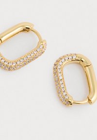 Złote kolczyki w kształcie owalnym, ozdobione przezroczystymi cyrkoniami po jednej stronie, charakteryzujące się polerowanym wykończeniem i gładką fakturą.