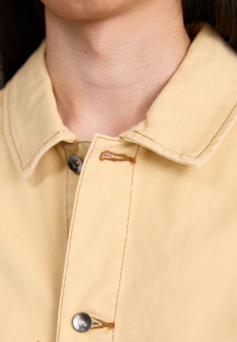 Camisa beige de botones con cuello, costuras en contraste y un detalle de botón metálico cerca del área del cuello. Tela suave y texturizada.