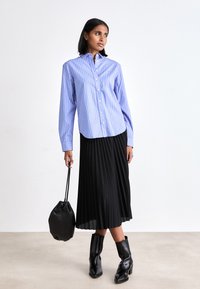 Blauw en wit gestreepte overhemd met knopen, lange mouwen, gecombineerd met een zwarte plissé rok en zwarte haklaarzen, met een zwarte tas in de hand.