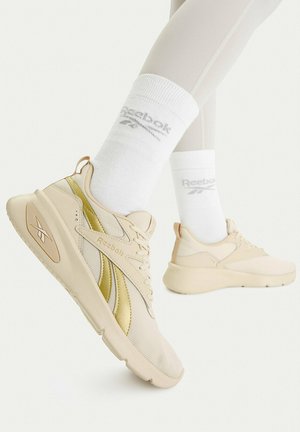 Beine mit weißen Reebok-Socken und beigefarbenen Reebok-Sneakern mit goldenen Akzenten vor weißem Hintergrund, Seiten- und Rückansichten gezeigt.
