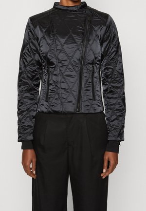 Personne portant une veste matelassée noire avec une fermeture éclair frontale asymétrique et des poches latérales zippées, associée à un pantalon noir ample.