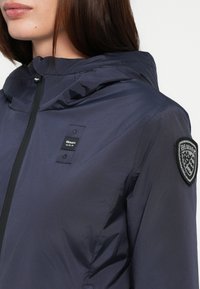 Marine waterdichte jas met hoge kraag, ritssluiting aan de voorkant en een merkpatch op de linker mouw. Glad textuur en lichtgewicht ontwerp.