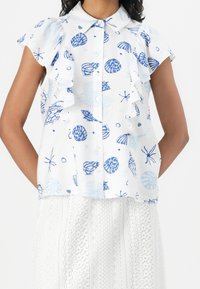 Blusa bianca senza maniche con motivi di conchiglie blu e dettagli arricciati, abbinata a una gonna bianca in pizzo.