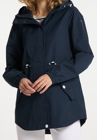 Mujer con chaqueta con capucha de color azul marino, con puños elásticos y bolsillos frontales, ajustando el dobladillo de la chaqueta, con pantalones blancos visibles.