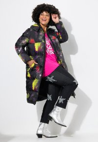 Cappotto puffer con base nera, design a macchie di vernice multicolore; lungo, con zip e tasche laterali. Indossato sopra una maglietta rosa e jeans strappati.