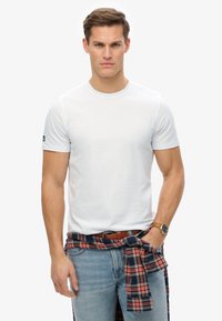 Hvid bomulds T-shirt med korte ærmer, rund halsudskæring og logo-detalje på ærmet. Båret med blå jeans og en ternet skjorte bundet om taljen.