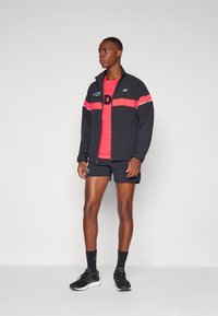 Black athletic jacket with red and white horizontal stripes, red t-shirt underneath, black shorts, and black running shoes.