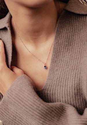 Silberne Halskette mit einem Tropfen-Anhänger, der einen zentralen Smaragd und rosafarbene Edelsteine zeigt, getragen auf einem nackten Schlüsselbein, vor einem beigen Pullover.