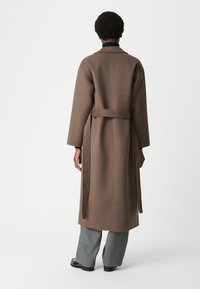Lungo cappotto di lana marrone con vita con cintura, maniche ampie e colletto arrotondato, abbinato a pantaloni grigi a gamba larga e scarpe nere.