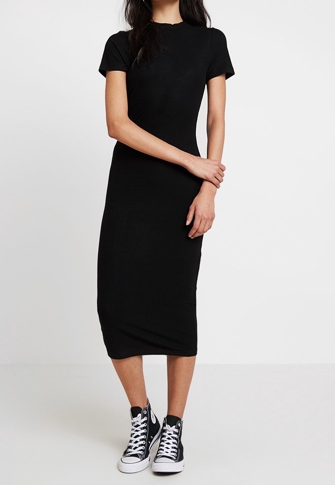 Femme portant une robe midi noire ajustée à manches courtes, associée à des baskets montantes noires, debout devant un fond blanc.