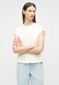 Mujer con cabello largo y castaño, que lleva una blusa blanca de manga corta con cintura fruncida y jeans azul claro, con los brazos cruzados, mirando de lado.