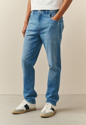Osoba nosząca jasnoniebieskie spodnie jeans o prostych nogawkach, białe sportowe buty z czarnymi paskami i białą koszulę, z jedną ręką w kieszeni.