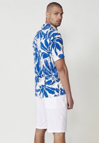 Cămașă cu mânecă scurtă de culoare albă, cu un model floral albastru, asortată cu o pereche de pantaloni scurți albi.  Material neted, cu o croială lejeră și design cu guler.