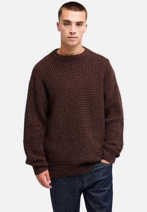 Junger Mann trägt einen dunkelbraunen, strukturierten Strickpullover und blaue Jeans, steht mit einer Hand in der Tasche vor einem einfarbigen hellen Hintergrund.