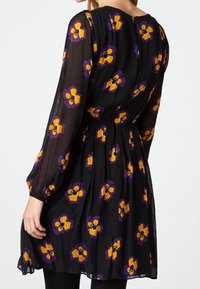 Robe noire transparente avec des manches longues, ornée de motifs floraux vibrants jaunes et violets, cintrée à la taille et évasée au bas.