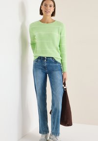 Pull côtelé vert clair avec des rayures blanches, jean bleu et baskets grises. Tenant un sac fourre-tout en daim marron. Tenue simple et décontractée.