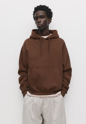 Braune Baumwollkapuzenjacke mit einer Fronttasche, Kordelzugkapuze und lässiger Passform, kombiniert mit hellgrauen, gefalteten Hosen.