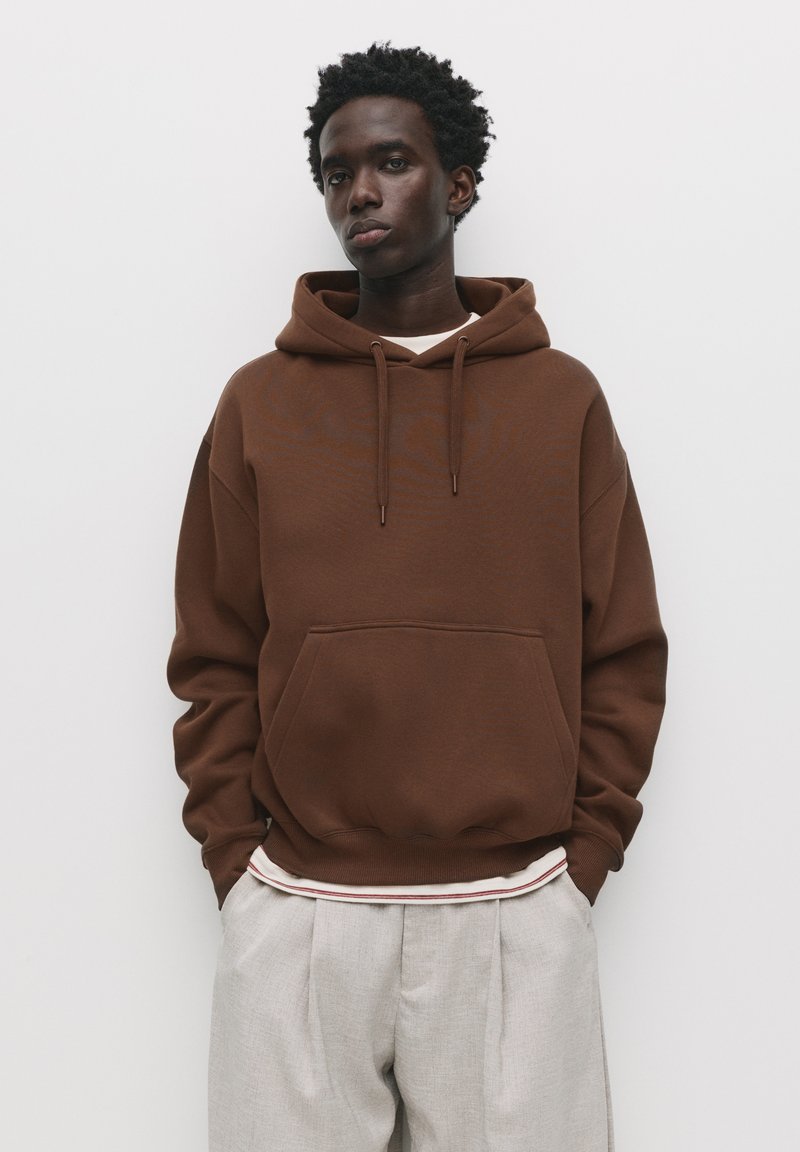 Brown cotton hoodie with a front pocket, drawstring hood, and relaxed fit, paired with light gray pleated trousers.