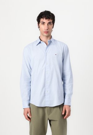 Lyseblå button-up skjorte med et lille prikmønster, krave og lange ærmer. Diskret logoaccent på brystet. Materialet er bomuld.