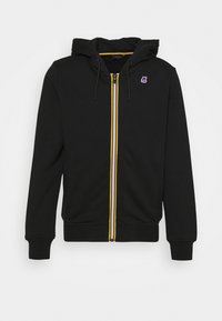 Haut à fermeture éclair noir en coton avec une capuche doublée. Présente des rayures contrastées jaunes, blanches et rouges le long de la fermeture éclair et un patch logo.