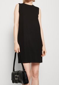 Abito nero a coste con scollo rotondo e senza maniche. La modella tiene in mano una borsa nera con una chiusura rettangolare.