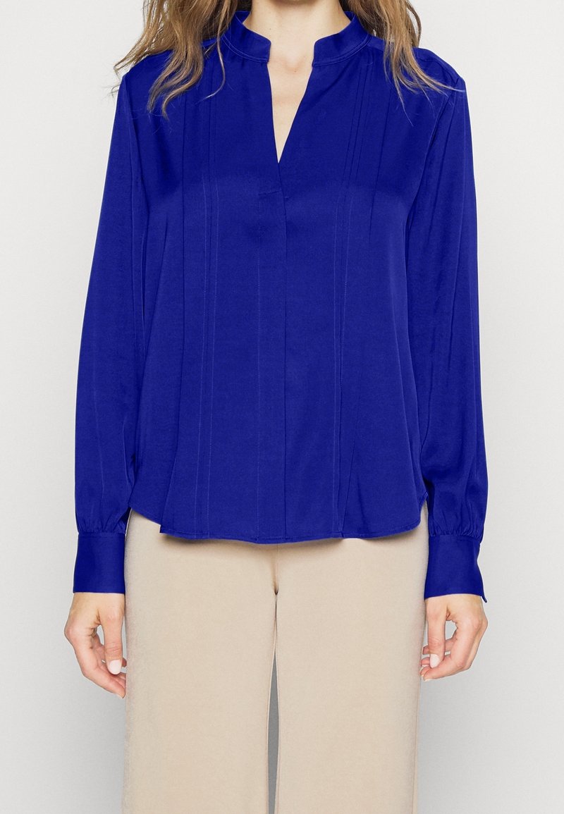 Femme portant un chemisier bleu roi à manches longues avec encolure fendue et un pantalon beige, montrée des épaules aux cuisses, debout devant un fond uni.