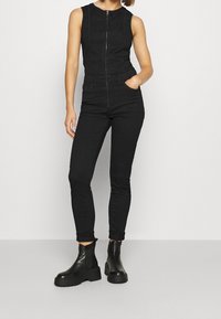 Czarny bezrękawnik z zamkiem błyskawicznym z przodu, w połączeniu z czarnymi dopasowanymi dżinsami i masywnymi czarnymi botkami za kostkę. Gładki, elegancki materiał i dopasowany krój.