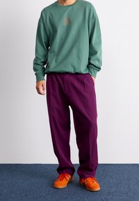 Zielony sweatshirt z okrągłym dekoltem i subtelnym nadrukiem graficznym, zestawiony z luźnymi fioletowymi spodniami i jasnopomarańczowymi tenisówkami.