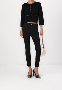 Black cropped sweater with silver stud accents, paired with tailored black pants and pointed black heels. Beige handbag in hand.