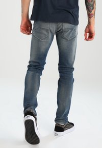 Jeans ajustados de color azul desgastado con cinco bolsillos, ligeros desgastes y una textura suave, combinados con zapatos de slip-on negros con estampado.