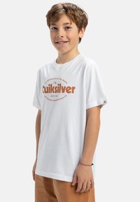 Bílá bavlněná tričko s krátkými rukávy, s velkým oranžovým logem "Quiksilver" a kulatým výstřihem, nošené s hnědými kalhotami.