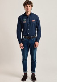 Camisa de botones color navy con logo y número 21, combinada con vaqueros ajustados y zapatos de ante marrón oscuro. Cuello contrastante y puños doblados.