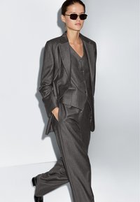 Kobieta idąca w ciemnoszarym garniturze w prążki z kamizelką, z rękami w kieszeniach, nosząca czarne okulary przeciwsłoneczne i czarne buty na tle jednolitego jasnego tła.