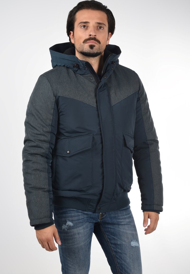 Giacca blu navy e grigia con cappuccio, due tasche frontali e un mix di materiali lisci e testurizzati, con un design aderente.