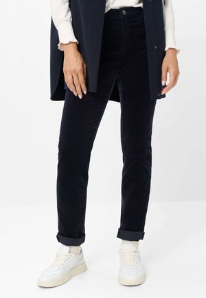 Person iført opkørte sorte bukser, hvide sneakers, cremefarvede sokker, en hvid langærmet top og en mørk vest mod en hvid baggrund.