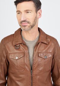 Braune Lederjacke mit einem geknöpften Kragen, Reißverschluss vorne und zwei Brusttaschen. Mit gesteppten Details an den Schultern und glatter Oberfläche.