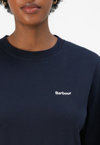 Σκούρο μπλε φούτερ με στρογγυλή λαιμόκοψη, διαθέτοντας το λευκό λογότυπο "Barbour" στην αριστερή πλευρά. Το ύφασμα φαίνεται απαλό και λείο.