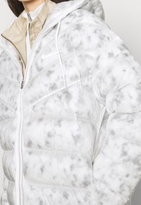 Vit puffjacka med kviltad design och en subtil svart marmormönster, med dragkedja fram och huva med justerbara snoddar.