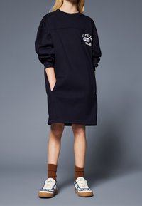 Námořnická mikinová šaty s dlouhými rukávy, s logem na hrudi, předními kapsami a volným střihem, doplněné hnědými ponožkami a vzorovanými botami.