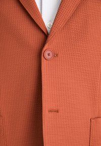 Rustfärgad texturerad blazer med snedskärning, en rund knappslutning och en sydd fickdetalj.