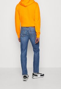 Orange kortärmad hoodie, blå denimjeans med en subtil tvätt och svarta och vita sneakers. Utsikt bakifrån som framhäver passform och färger.
