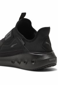 Scarpa sportiva nera con tomaia in tessuto testurizzato, suola in gomma e imbottitura visibile. Presenta un manico e dettagli di branding.