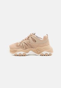 Scarpe da ginnastica beige con tomaia in rete e suede, suola bianca testurizzata, design ondulato unico e dettaglio di branding minimalista sul lato.