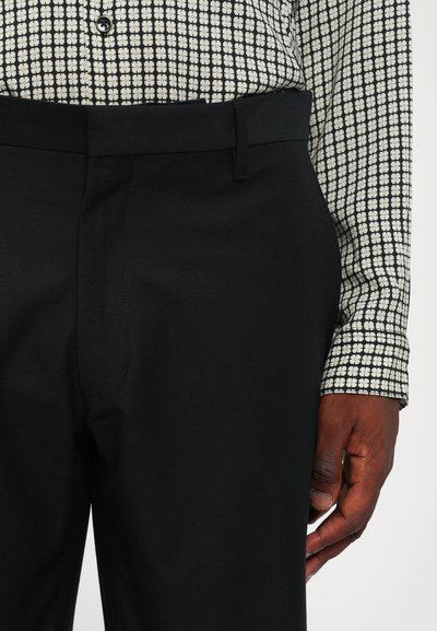 Pantalons noirs en tissu lisse, avec un devant plat et des passants de ceinture, associés à une chemise à manches longues à motifs noir et blanc.