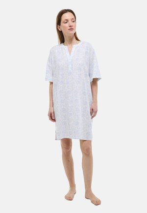 Femme debout pieds nus portant une robe fleurie bleu clair jusqu'aux genoux, à manches courtes et col en V, regardant légèrement vers la droite.