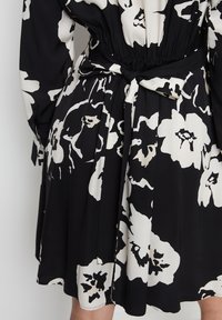 Schwarz Kleid mit großen weißen Blumenmustern, elastischem Bund mit gebundenem Gürtel, langen Ärmeln und einem ausgestellten Rock. Glatte Stoffoberfläche.